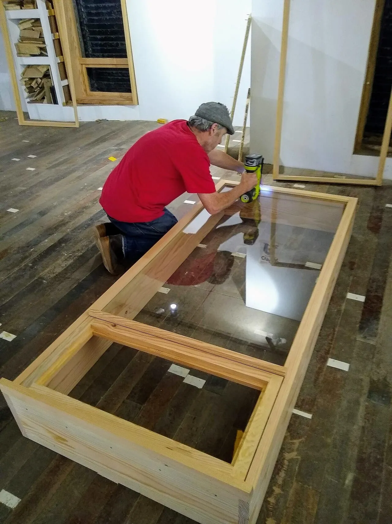 Menuisier agenouillé près d'un châssis de fenêtre en bois vitré posé au sol dans un atelier