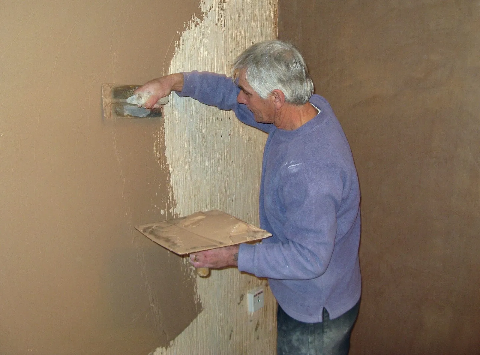 Plâtrier appliquant du plâtre à la truelle sur un mur intérieur, platoir garni dans l'autre main, dans une pièce en travaux
